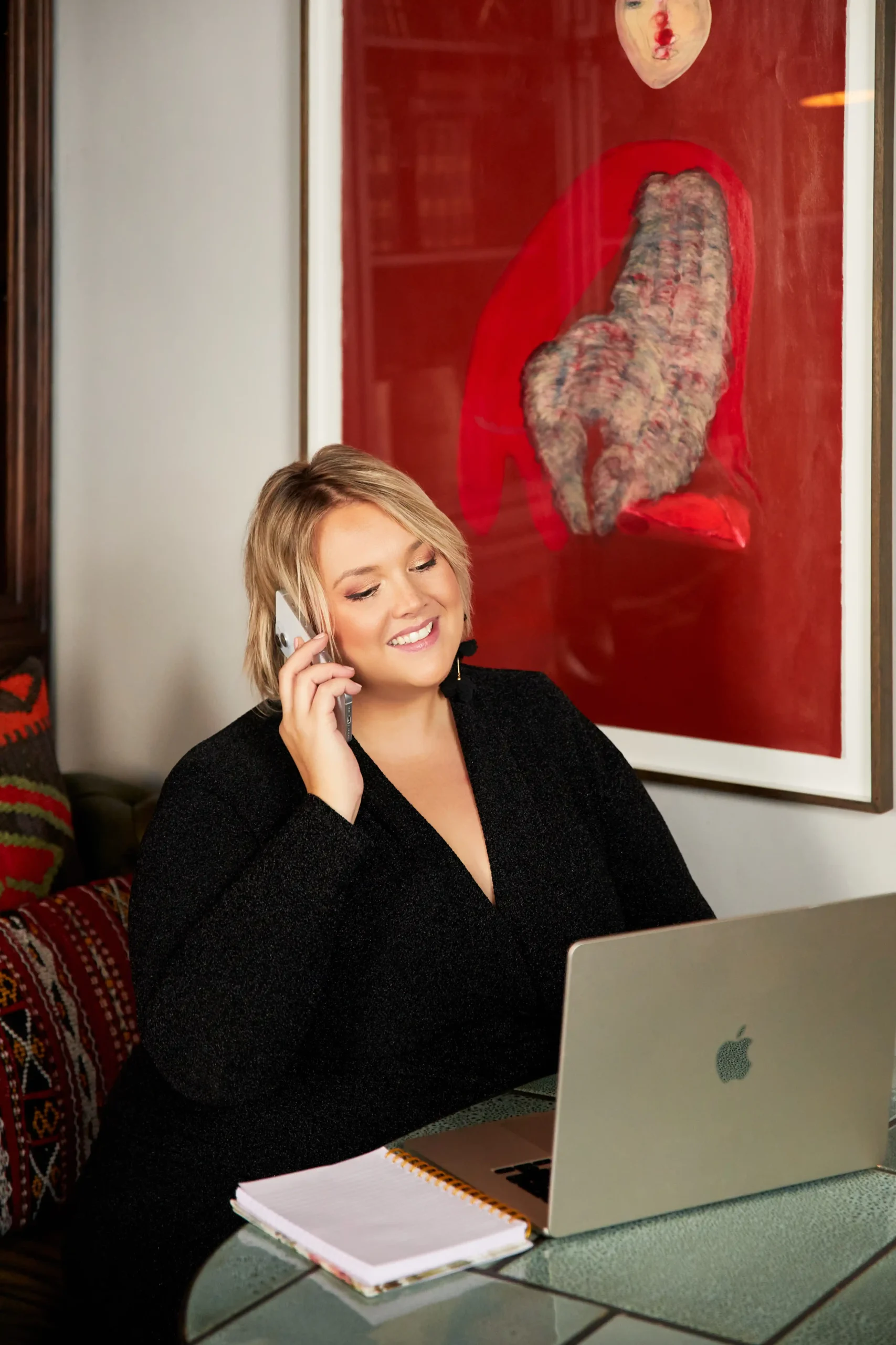 Smiling woman on phone with laptop and notebook in cozy workspace.